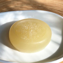 Load image into Gallery viewer, Closeup of a pale yellow, round, glycerin face soap with an embedded tree design, sitting on a white ceramic plate, on a blonde piece of wood, in dappled sunlight.
