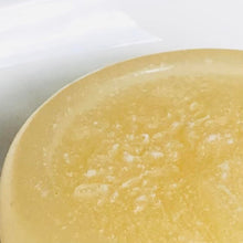 Load image into Gallery viewer, Closeup of the corner of a pale yellow, round, glycerin face soap with small, white specks of kaolin clay that can be seen, sitting on a white ceramic plate.