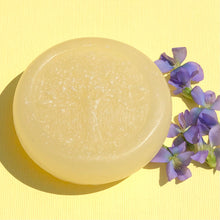 Load image into Gallery viewer, Closeup of a pale yellow, round, glycerin face soap with an embedded tree design, sitting on a bright yellow background, surrounded by small purple flowers on the right side.