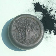 Load image into Gallery viewer, Closeup of a round, grey, glycerin face soap with an embossed tree design on the front and embedded black specks of charcoal that can be seen, laying on pale, aqua blue background, with a splash of charcoal pigment shown at the top right.