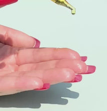 Load and play video in Gallery viewer, A closeup of a glass dropper tip full of golden oil falling in slow motion onto fingertips with long, red nails.
