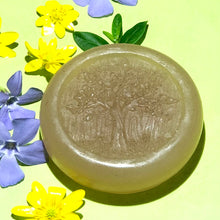 Charger l&#39;image dans la galerie, Closeup of a round, pale-colored, glycerin face soap with an embossed tree design on the front and embedded brown specks of rhassoul clay that can be seen,  laying on a bright yellow background with bright yellow and purple flowers, in bright sunlight. 
