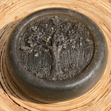 Charger l&#39;image dans la galerie, Closeup of a round, grey, glycerin face soap with an embossed tree design on the front and embedded black specks of charcoal and small, brown pieces of hemp that can be seen, laying against blonde wood in the sunlight. 
