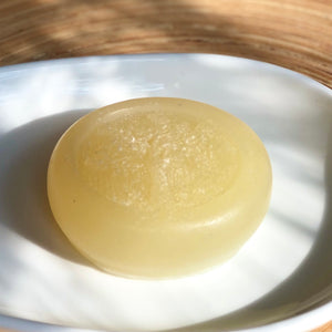 Closeup of a pale yellow, round, glycerin face soap with an embedded tree design, sitting on a white ceramic plate, on a blonde piece of wood, in dappled sunlight.