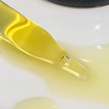 Load image into Gallery viewer, Extreme closeup of the tip of a glass dropper filled with shimmering, golden oil, laying in more golden oil on a shiny, white surface.
