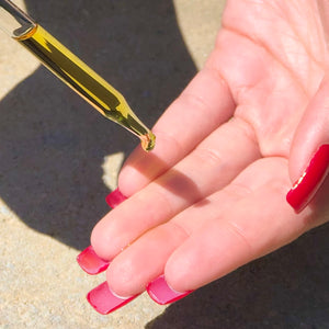 Closeup of a glass dropper of golden oil, with a drop about to fall onto fingertips sporting long, red nails.