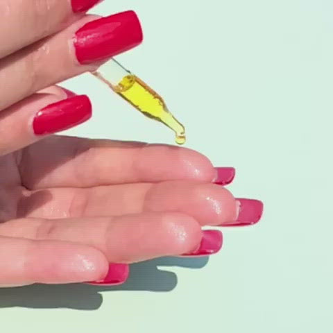A close-up drop of golden oil falls in slow motion from a glass dropper onto fingertips with long, red nails.