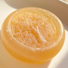 Charger l&#39;image dans la galerie, Closeup of a round, pale-colored, glycerin face soap with an embossed tree design on the front and embedded brown specks of rhassoul clay that can be seen, bathed in bright, dappled sunlight against a cream-colored background. 
